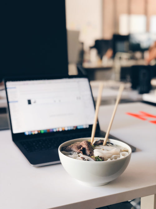 a bowl of noodles sits in front of a laptop on a desk