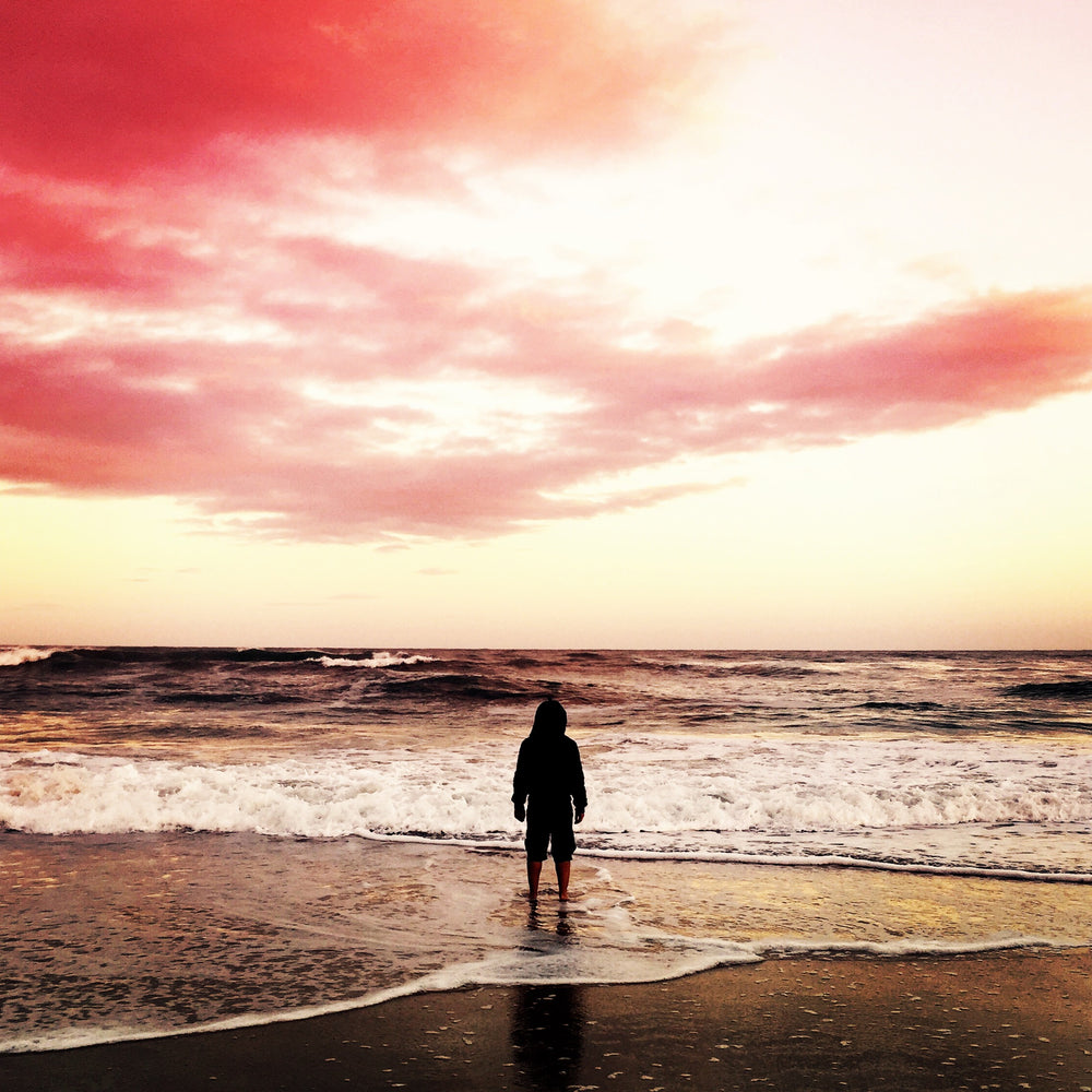 a figure in a hoodie on a beach at the shoreline