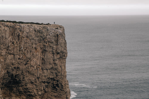 a figure on a cliff overlooking the ocean under a grey sky