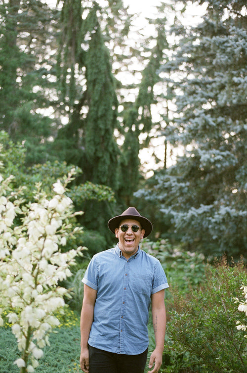 a man in a fedora and sunglasses beams in the woods