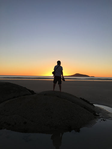 a man on a dune watches the sun set from the beach