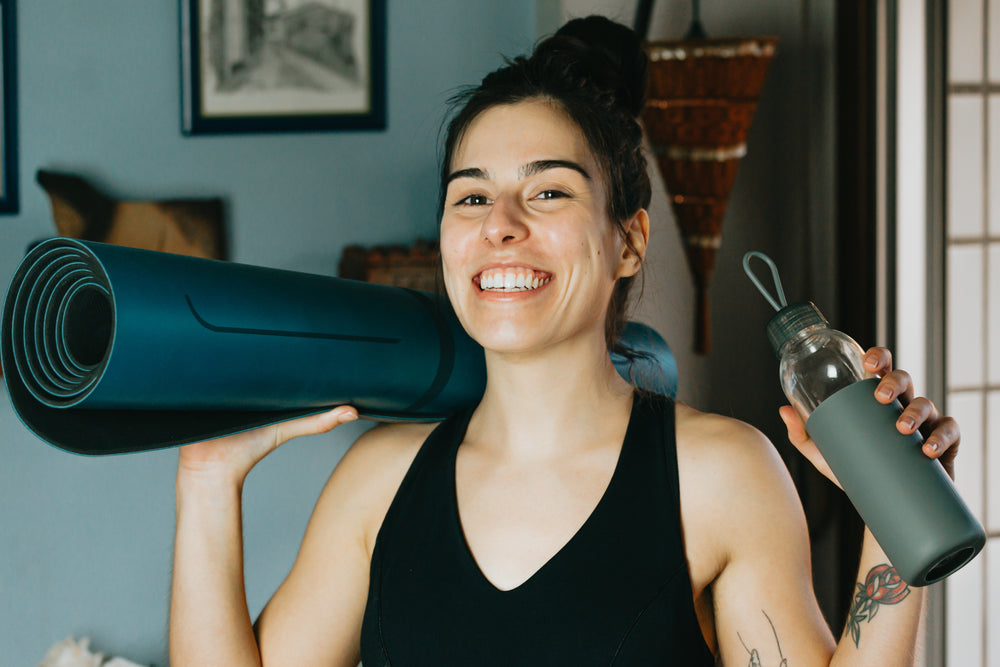 a person smiles holding a water bottle and a yoga mat