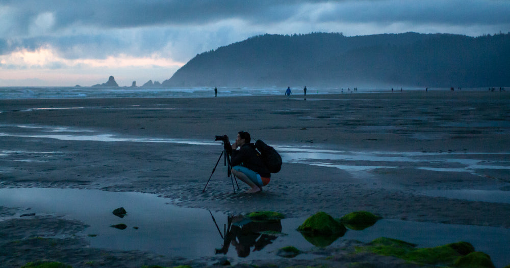 a photographer crouches at his tripod on a misty beach