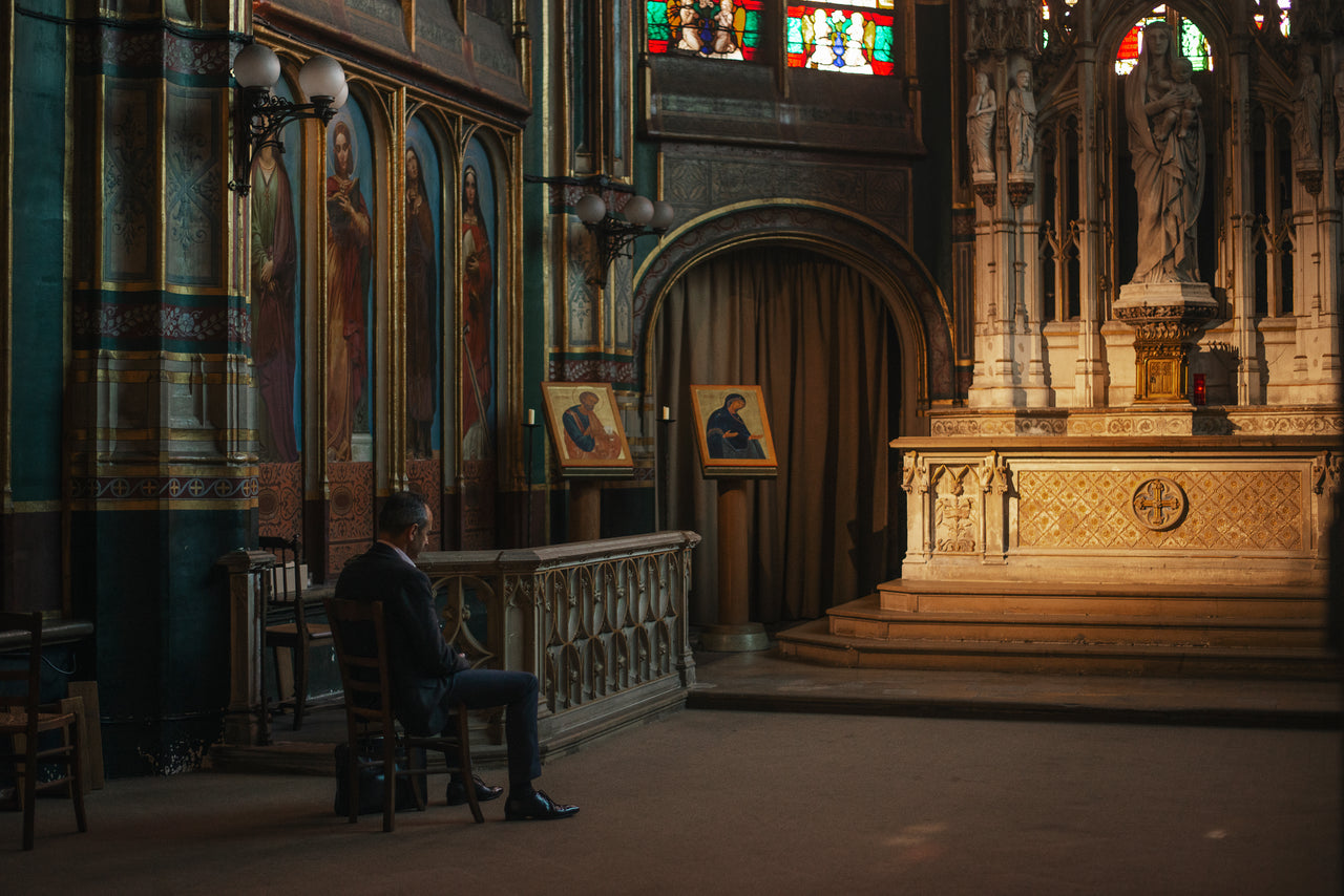 A man in a dark suit sitting alone on a wooden chair beside the chancel rail of an ornate church, with the lit stone altar to his right