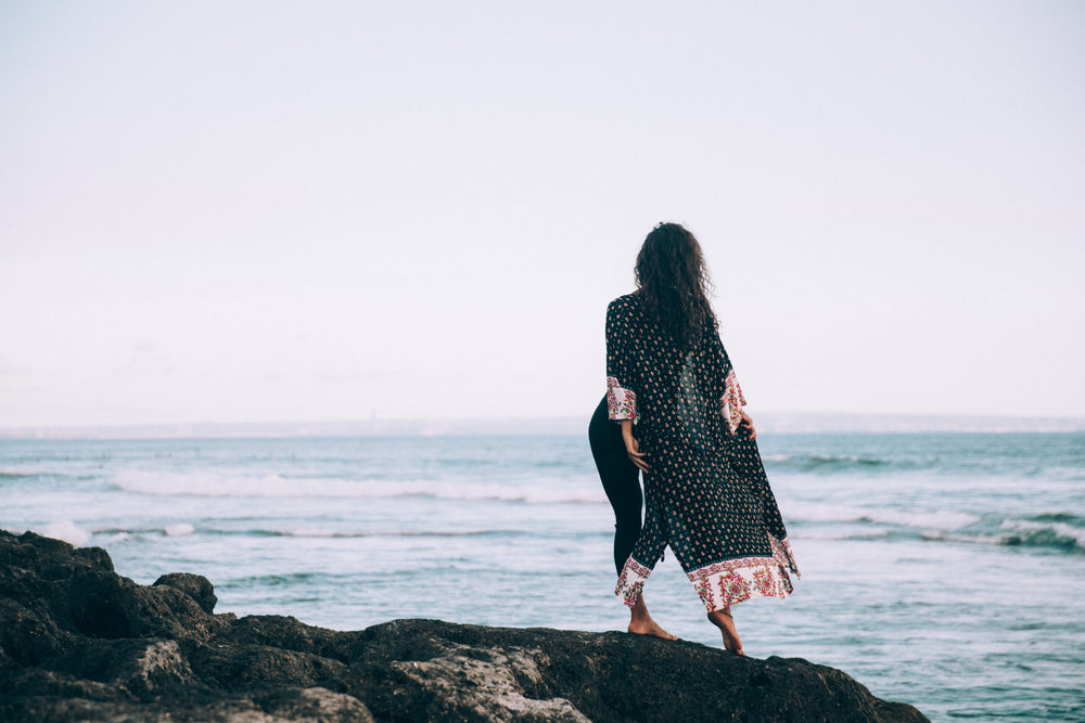 a woman in flowing patterned robe stands by waters edge