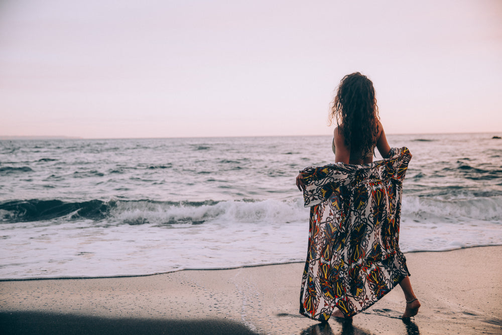 a woman on a beach opens her sarong at the rolling waves