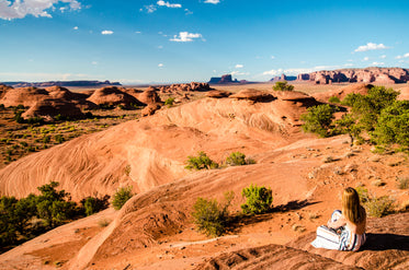 a woman sits on a desert hill overlooking mountain plains