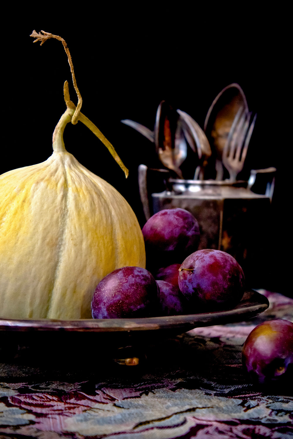 a wrinkly melon pushes plums out of a fruit bowl