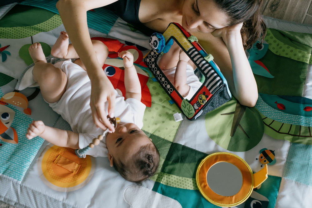 baby lays on a colorful mat with a person laying next to them