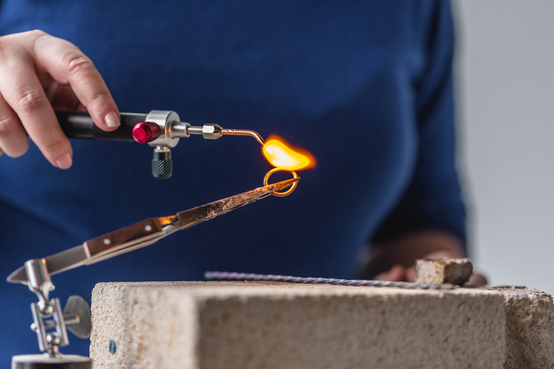 jeweler workshop scene with butane torch heating a small ring in tweezers on a firebrick block, illustrating the precision finishing behind a stepped wedding band