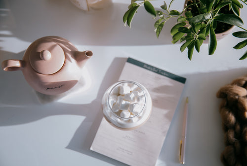 flatlay of pink teapot, paper, pen and a glass of marshmallows