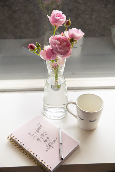 glass vase with pink flowers with notebook and a mug