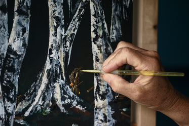 hand holds a paint brush to a painting of birch trees