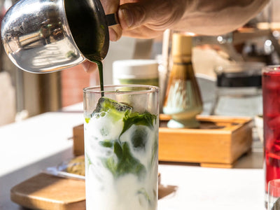 Hand Pours A Green Liquid Into A Glass Of Ice And Milk