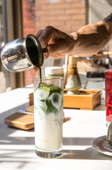 hand pours a green liquid into a glass of ice and milk