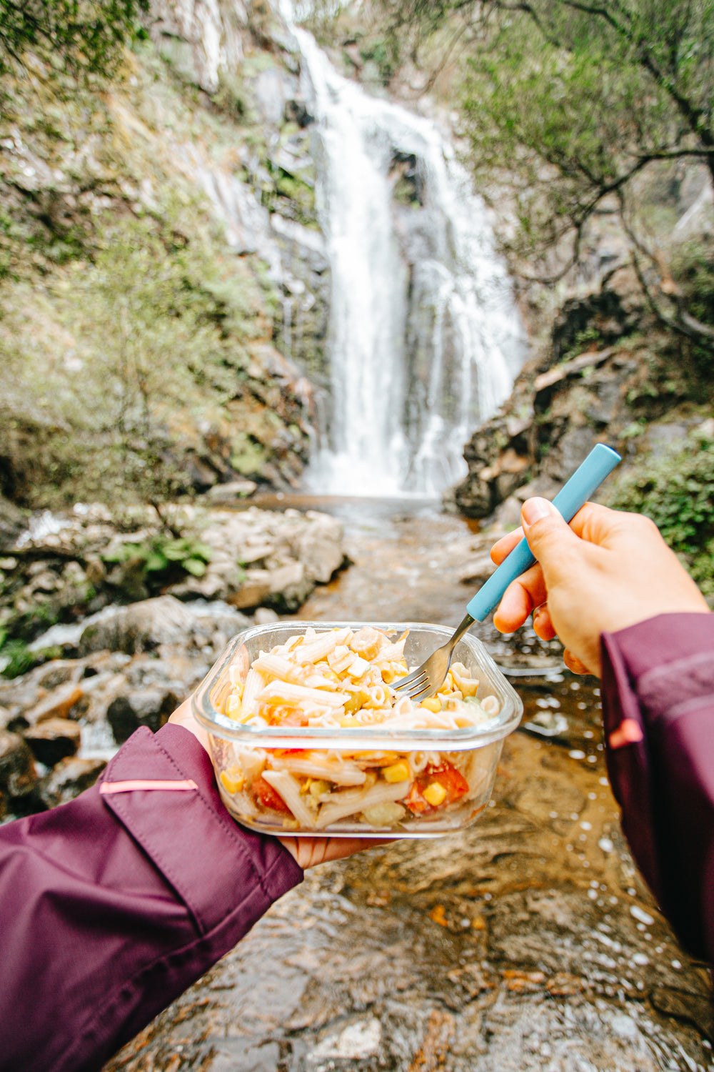 hands hold pasta salad and a fork by a waterfall outdoors