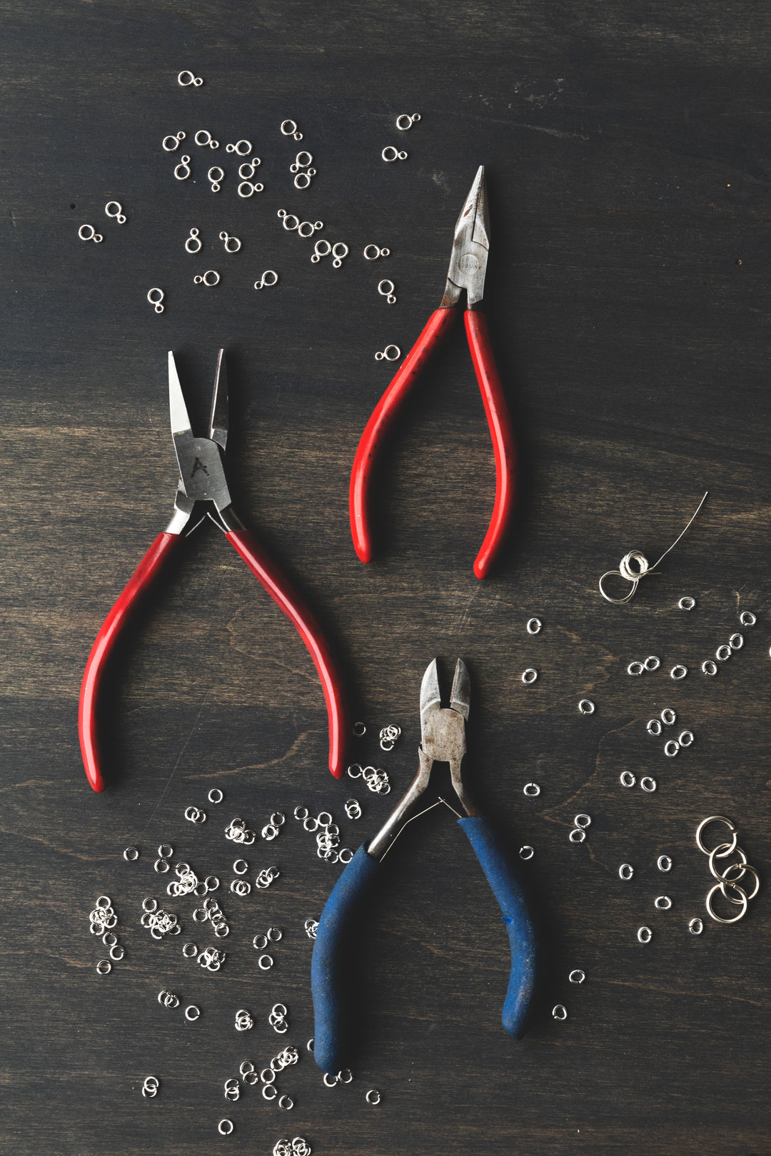 Three pairs of jeweler's pliers with red and blue handles laid on a dark wood bench with scattered silver jump rings around them