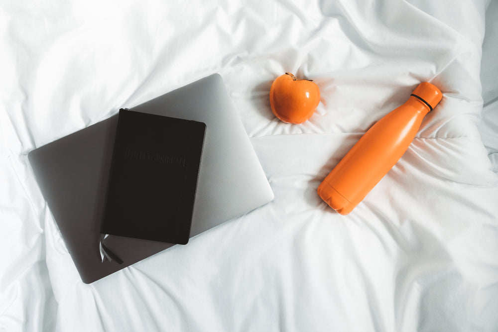 laptop with notebook and water bottle on white bed
