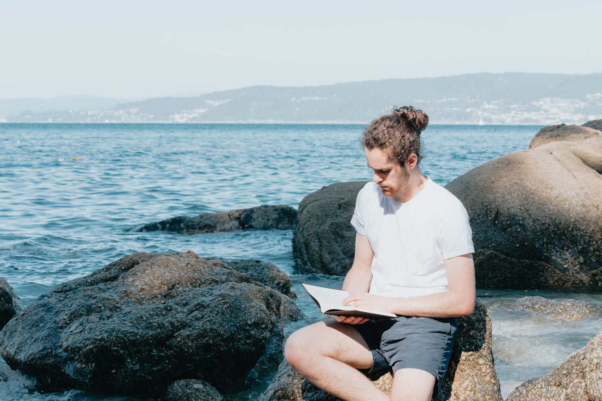 man sits on a rock by the water and reading his book