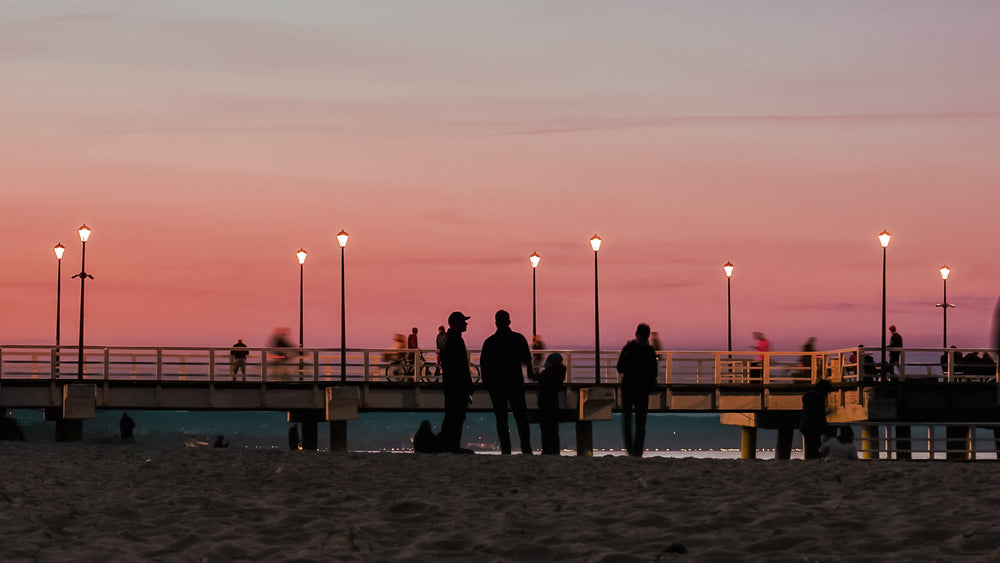 people stand on beach by a pier at sunset