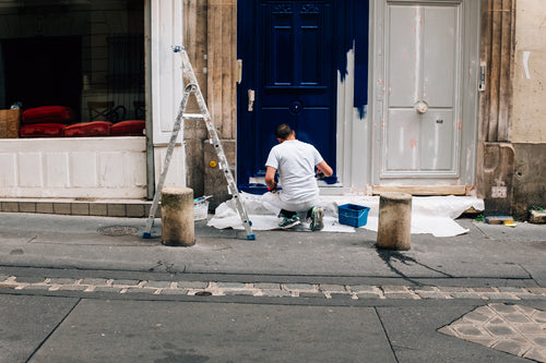 person crouches down on a street and paints a door white