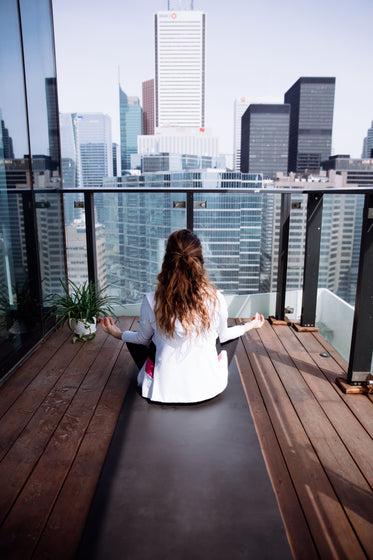 person faces the city while sitting on a yoga mat on a balcony