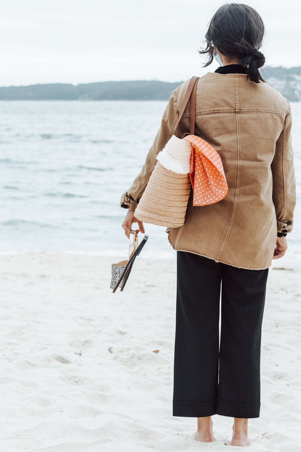 person faces the water and holds their shoes in hand