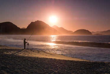 person fishing on the beach is silhouetted at sunset