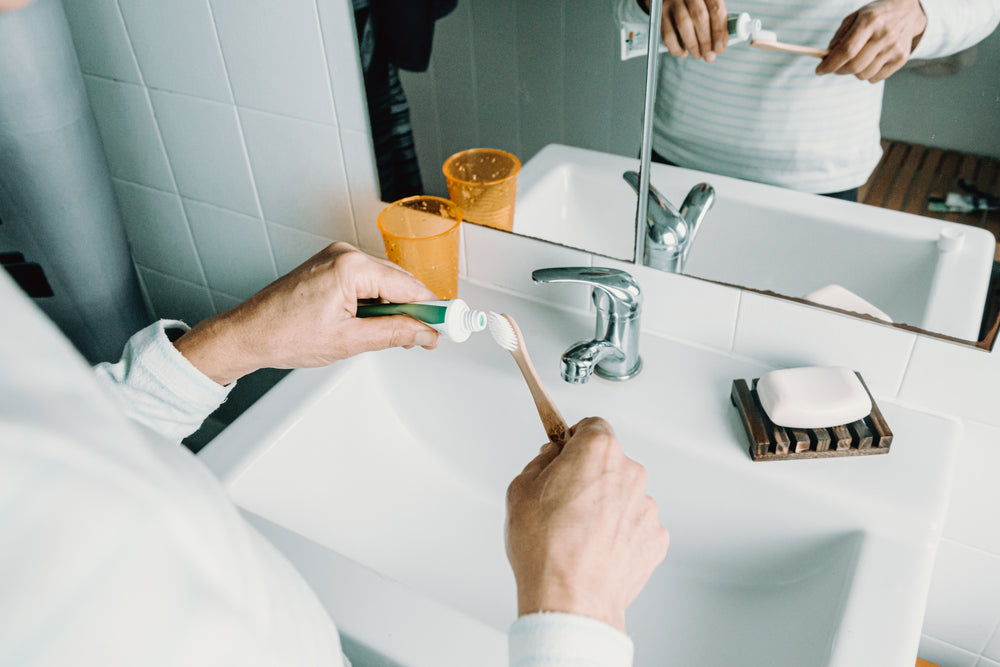 person holds a wooden toothbrush and toothpaste over a sink