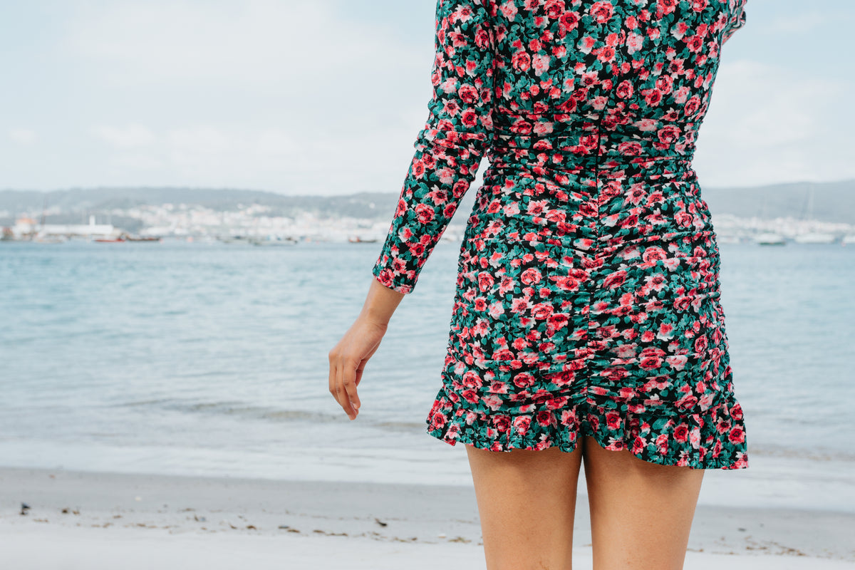 person in a floral dress stands on a beach facing the water