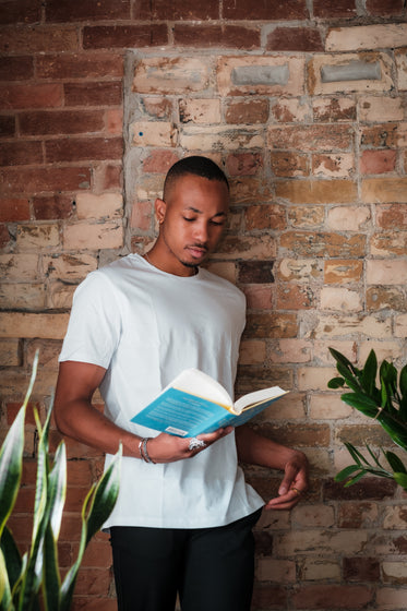 person in white stands reading a novel by a brick wall