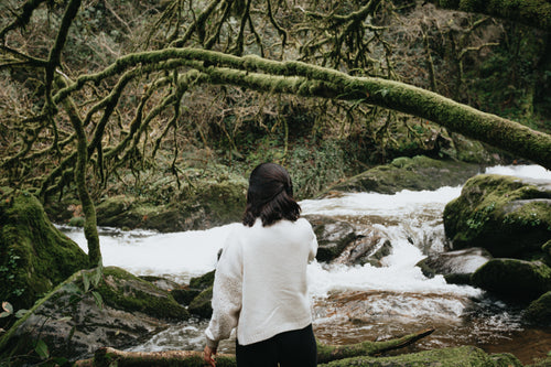 person in white sweater stands facing a rushing river