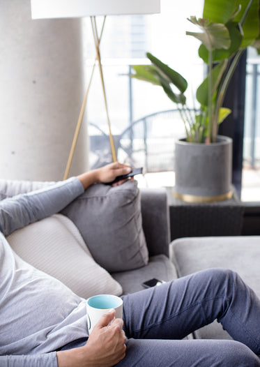 person leans back on a grey couch while holding a mug