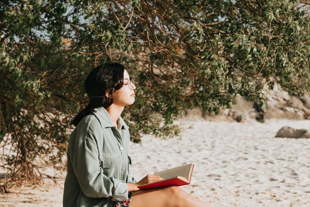 person reads a novel on the sandy beach by a tree