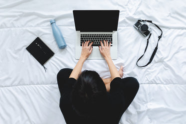 person sits cross legged on bed and types on their laptop