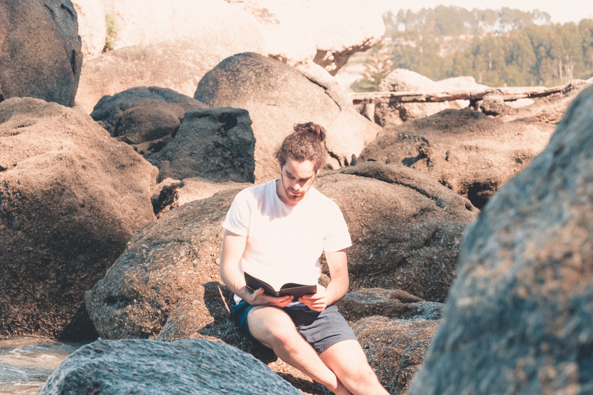 person sits on rocks and reads a novel in white shirt