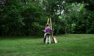 person sitting at an easel by green grass and trees