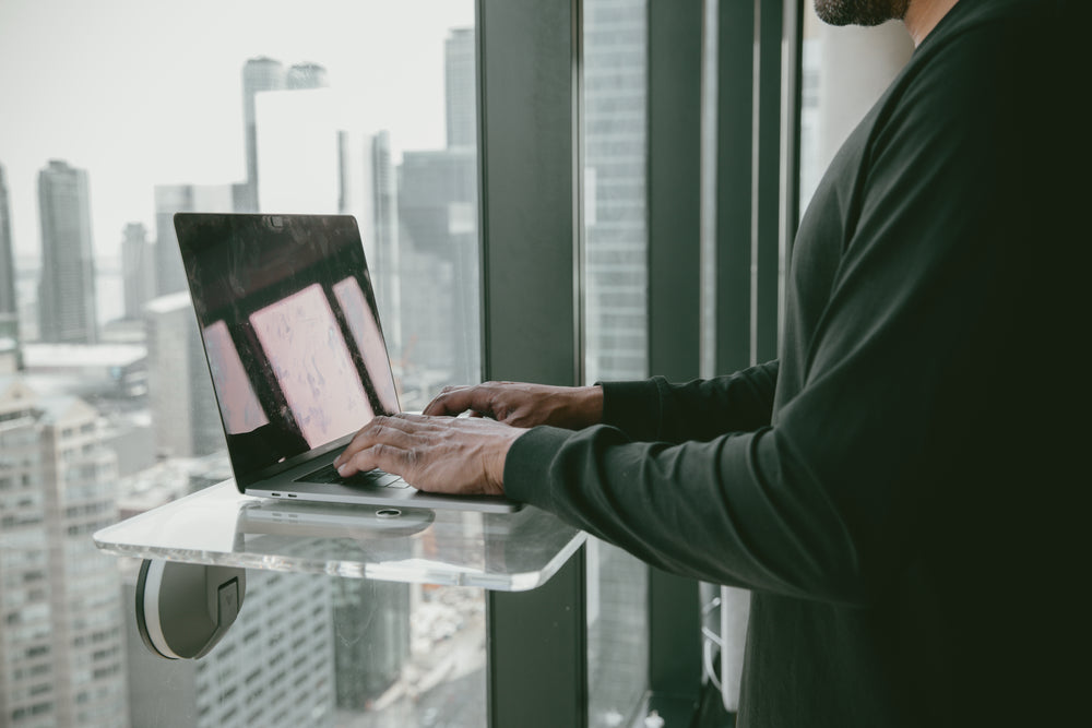 person stands and types on a laptop by a window