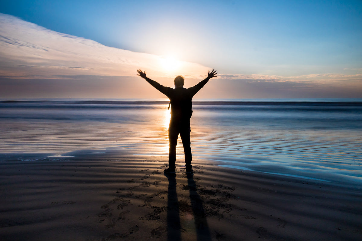 person stands on a beach with their arms out at sunset
