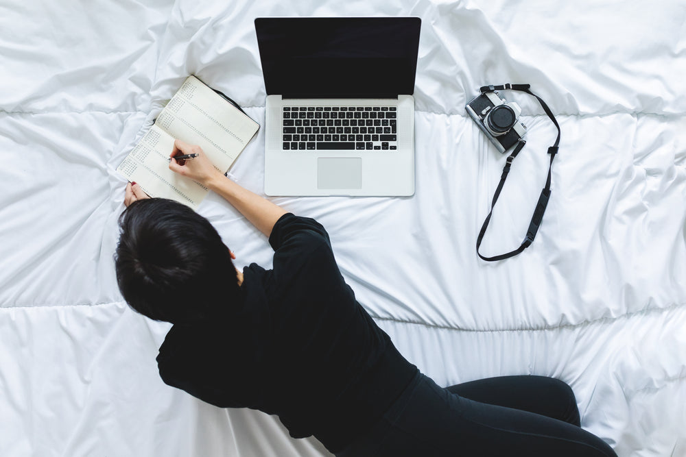 person writing in notebook beside laptop working from bed