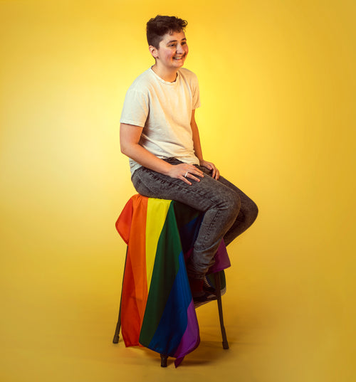 smiling trans man seated above draped rainbow flag