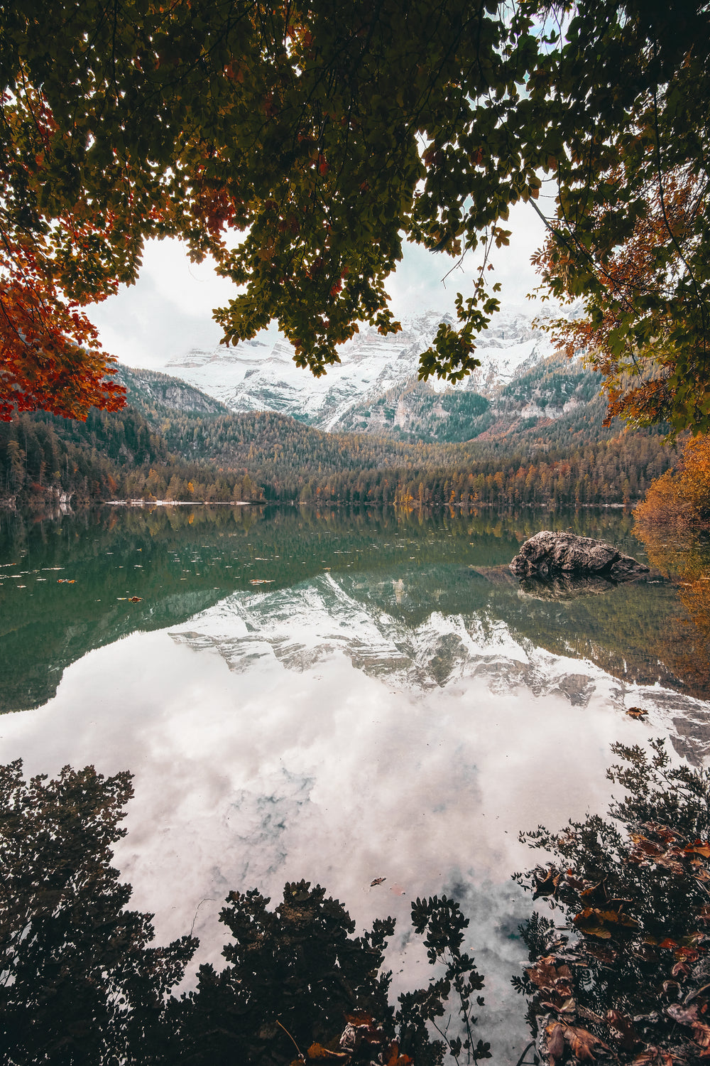 snow covered mountains reflect on the water by a fall forest