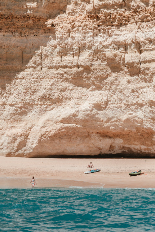 sunbathers, surfers and kayakers on a beach under cliffs