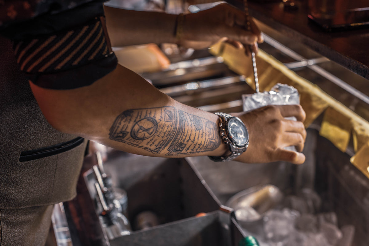 tattooed person holding a glass with ice over a bar well