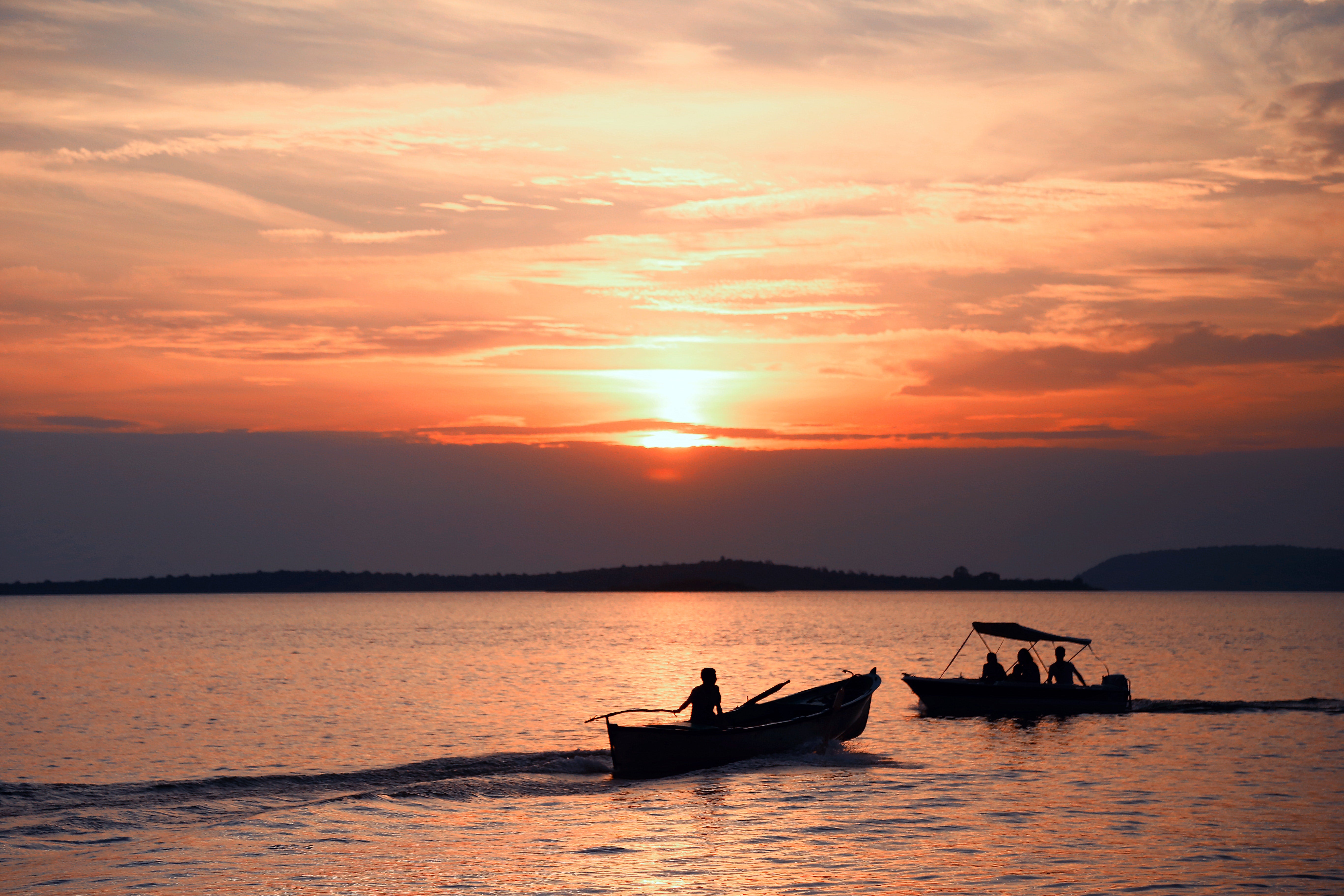 Two men fishing in boat sunset sky sea shore mountains - Stock