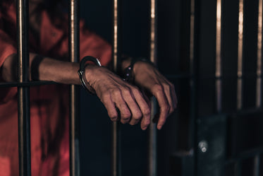 two hands in handcuffs lean on bars of a prison cell