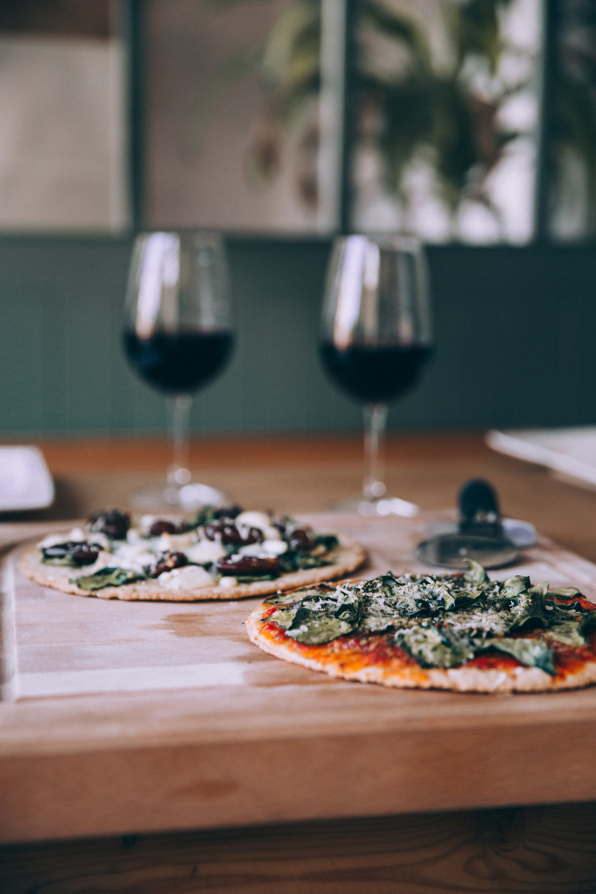 two large pizzas on wooden board with glasses of wine nearby