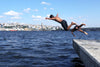 two people dive into blue water across from a cityscape