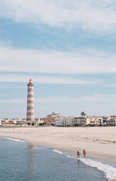 two people walk the beach with a lighthouse in the distance