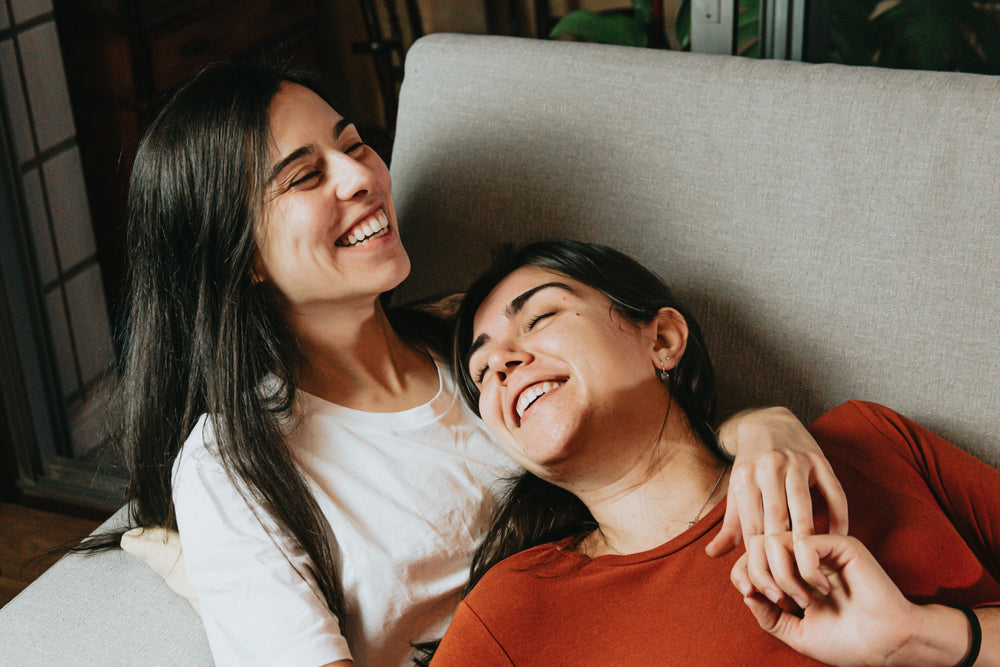 two woman smile wide and relax on a grey couch